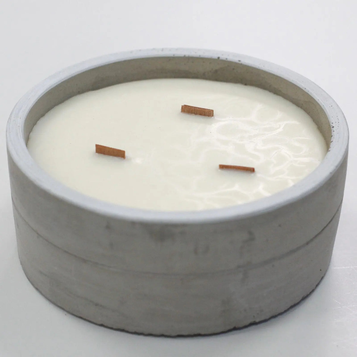 Vela redonda en recipiente de hormigón gris con tres mechas de madera y cera blanca sobre fondo neutro.