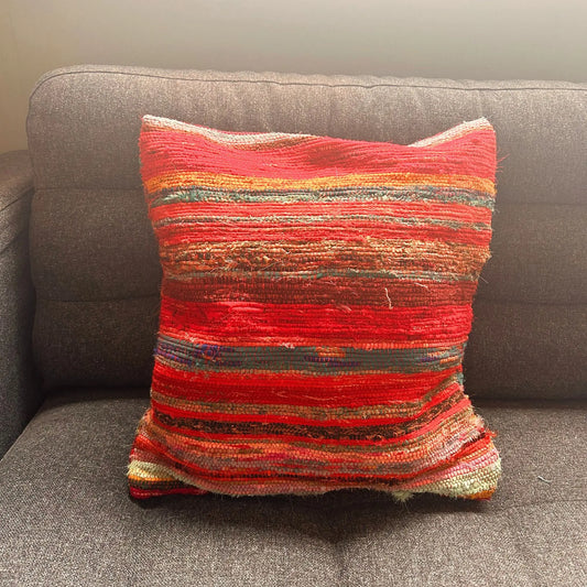 Funda de cojín artesanal de 40x40cm con diseño de rayas tejidas en tonos rojos y textura rústica sobre un sofá gris