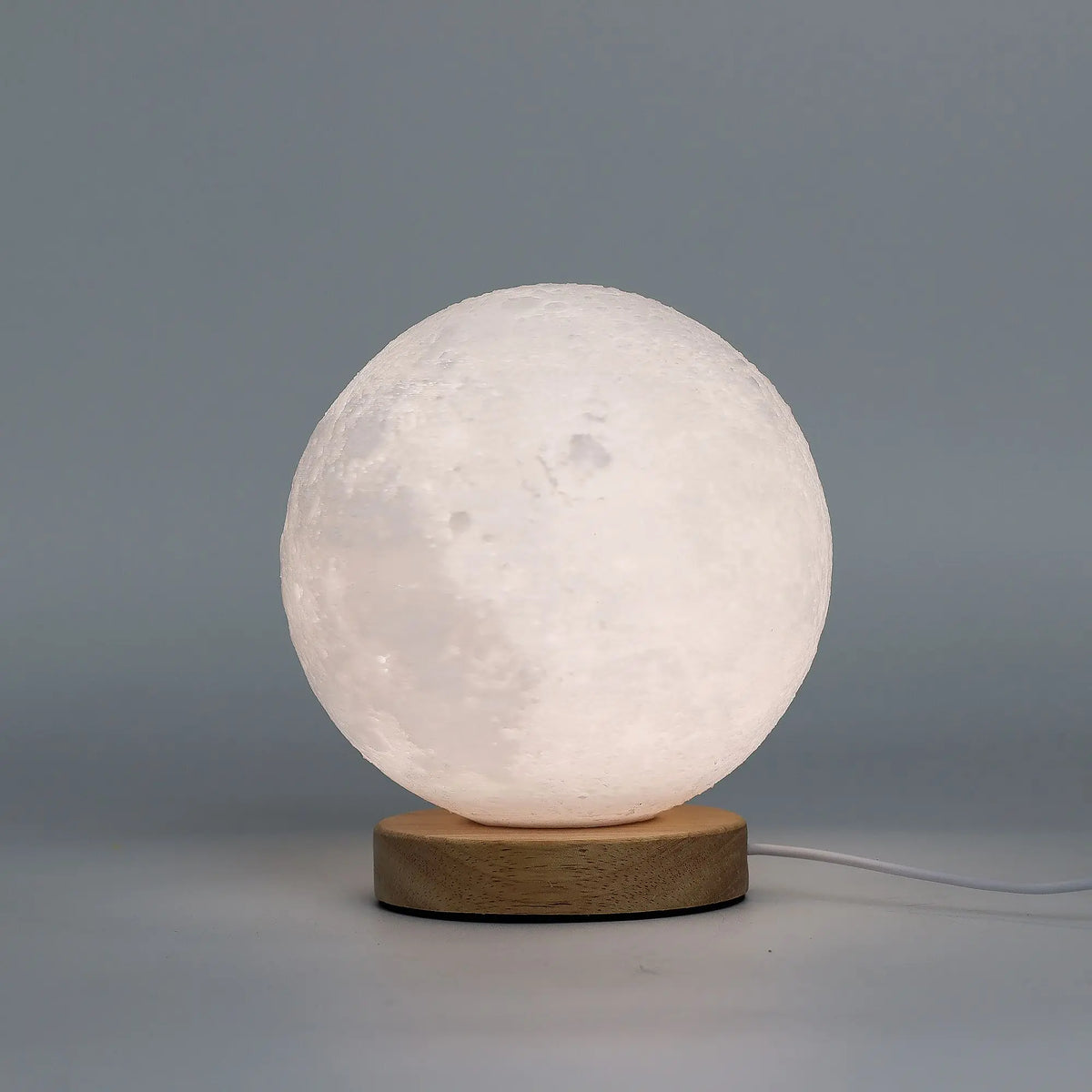 Lámpara esférica tipo luna con textura realista sobre base circular de madera clara, con cable USB, luz tenue cálida.