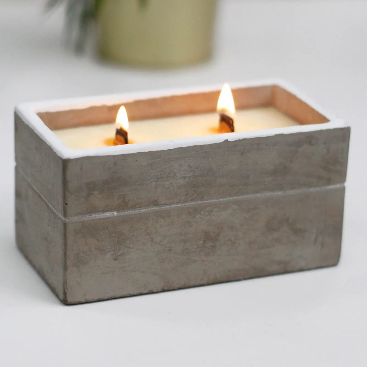 Vela rectangular de hormigón gris con mechas de madera encendidas en su interior sobre fondo blanco.
