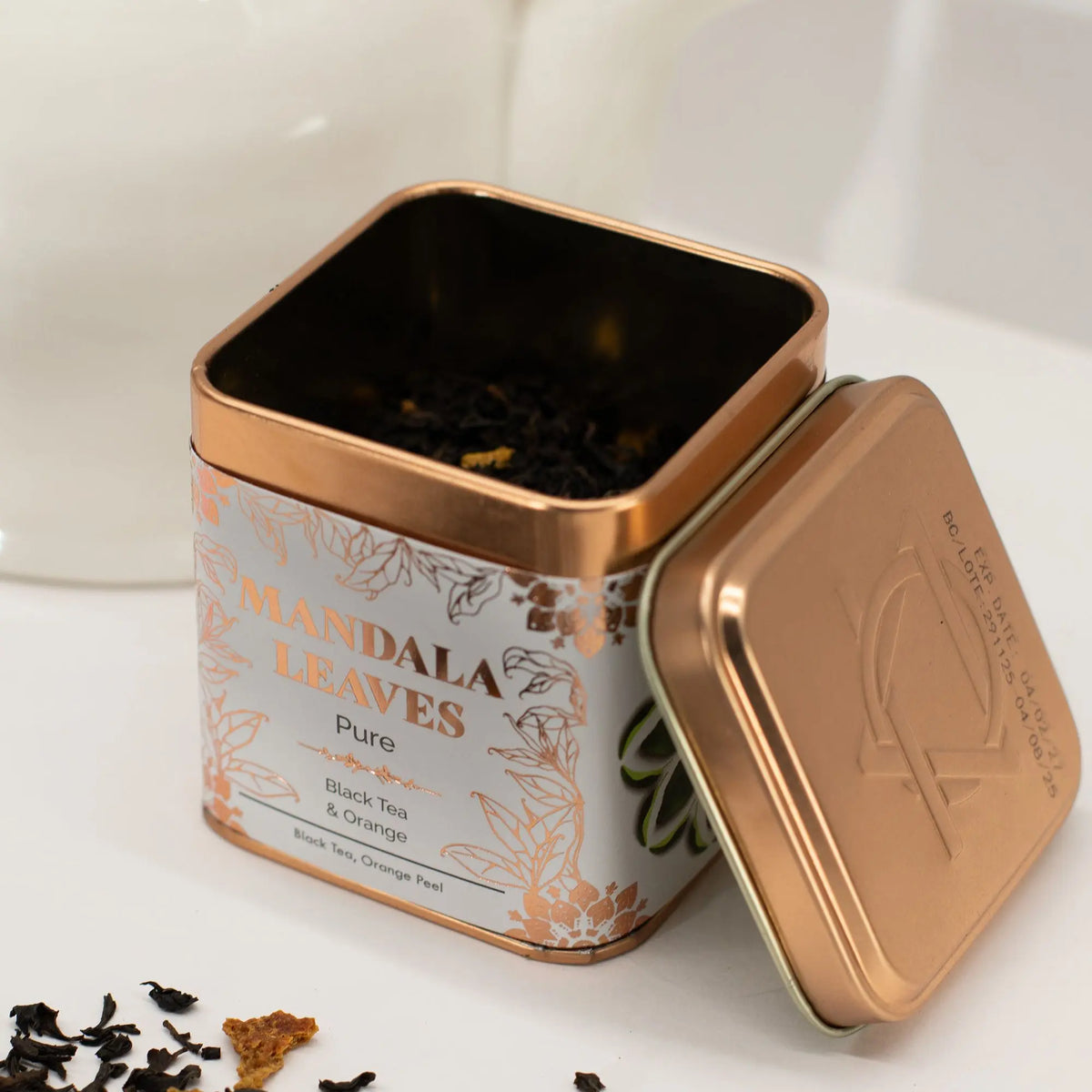Lata cuadrada de metal color cobre abierta, con hojas de té negro y trozos de naranja dentro sobre fondo blanco.