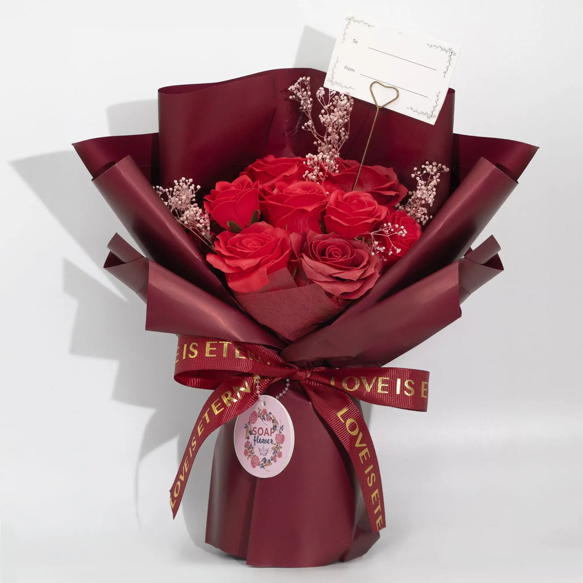 Ramo de rosas rojas en envoltura burdeos, con tarjeta y cinta dorada "Love Is Eternal". Ideal para regalos románticos.