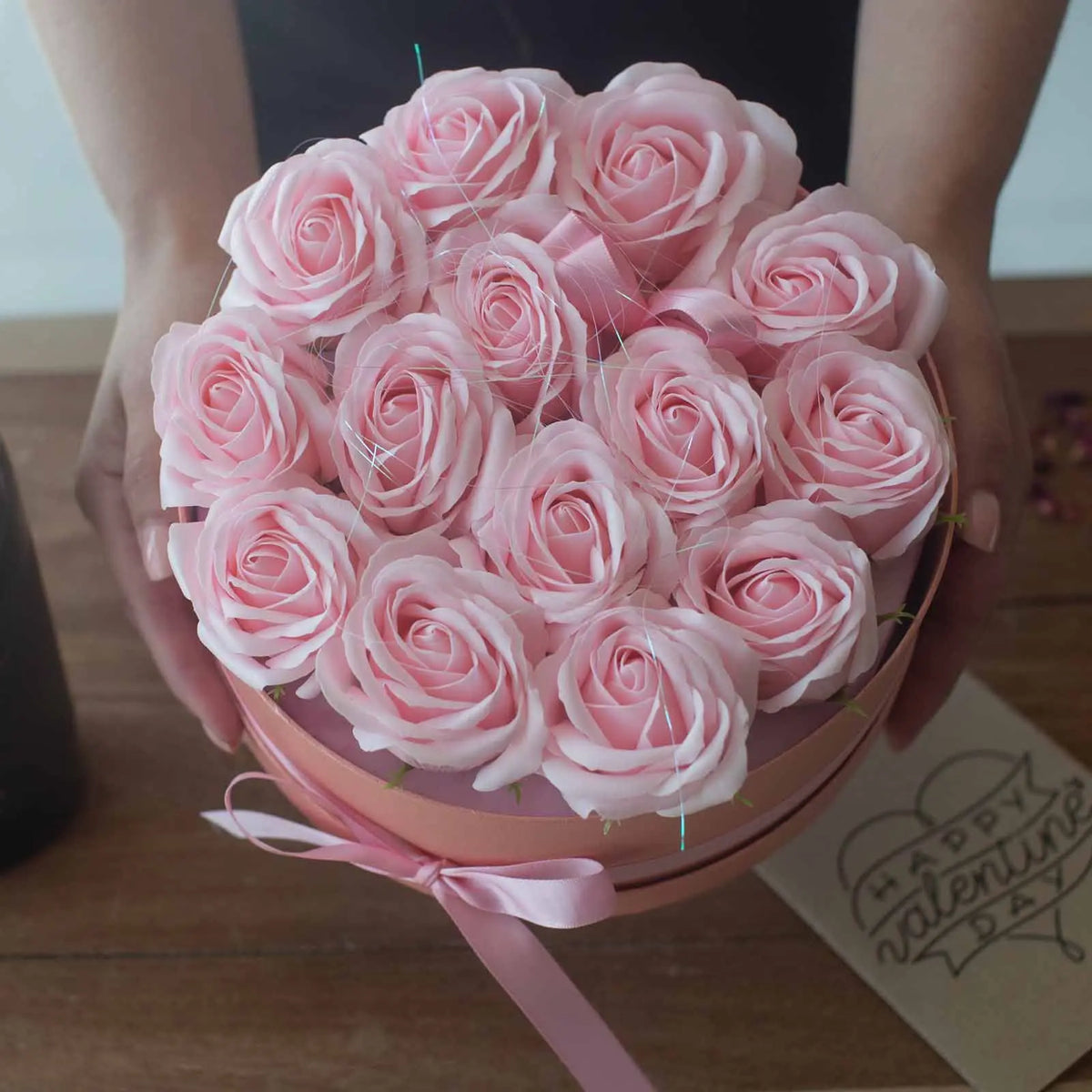Caja redonda rosa con 13 rosas de jabón en tonos rosados, decorada con un lazo, sostenida por manos.