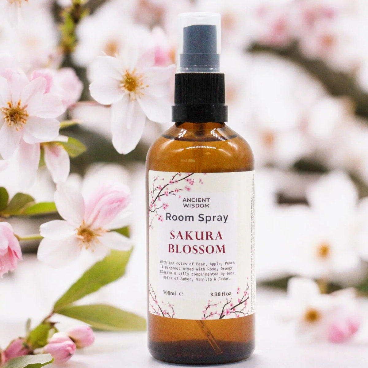 Spray de 100 ml en frasco de vidrio ámbar con atomizador negro, frente a flores de cerezo desenfocadas de fondo.