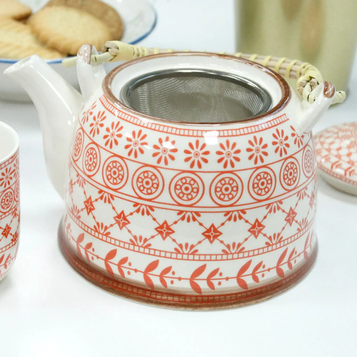 Tetera de cerámica blanca con diseño geométrico y floral naranja, asa de mimbre y colador de acero inoxidable incluido.