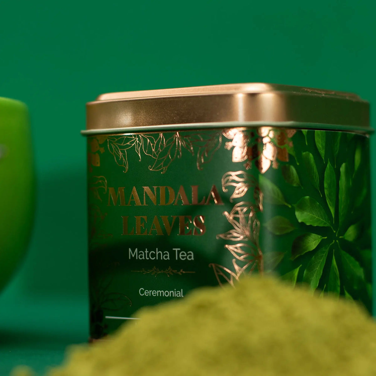 Lata cuadrada de té matcha verde con detalles dorados y tapa metálica sobre fondo verde con polvo de té al frente.
