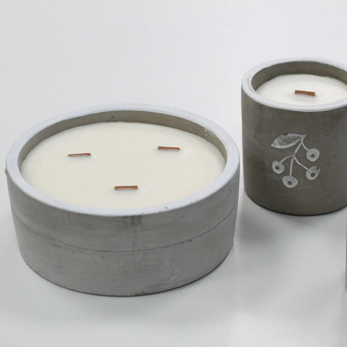 Velas de cera blanca en recipientes de hormigón gris con mechas de madera, sobre fondo blanco minimalista.