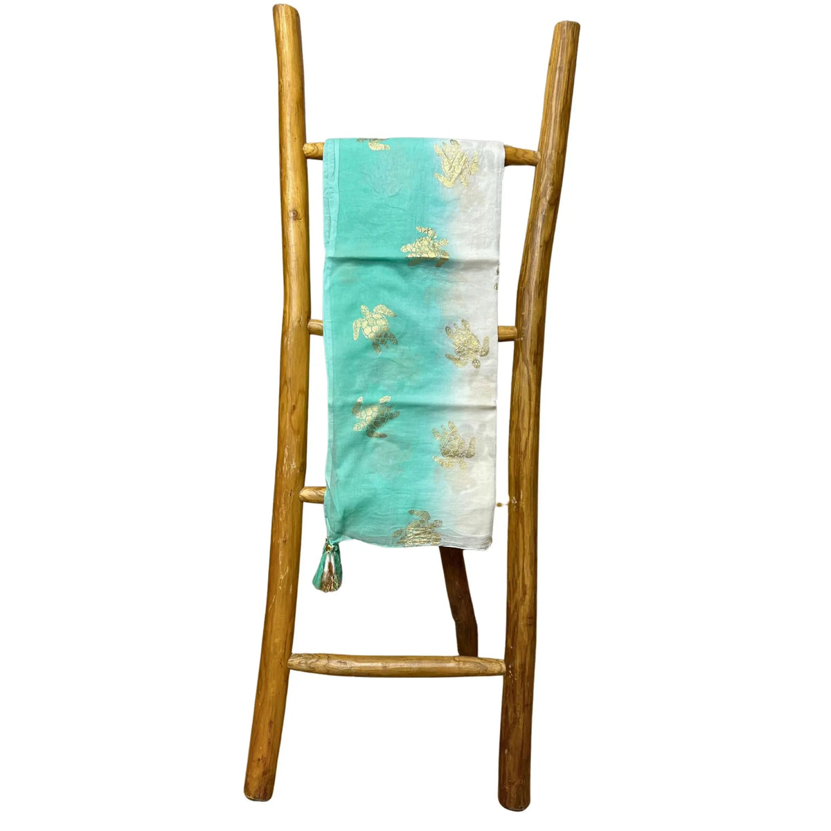Pareo de algodón blanco y turquesa con estampado de tortugas doradas, colgado en una escalera decorativa de madera.