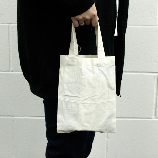 Bolso pequeño de algodón natural de 25x20 cm con asas cortas, sostenido por una persona con ropa oscura.