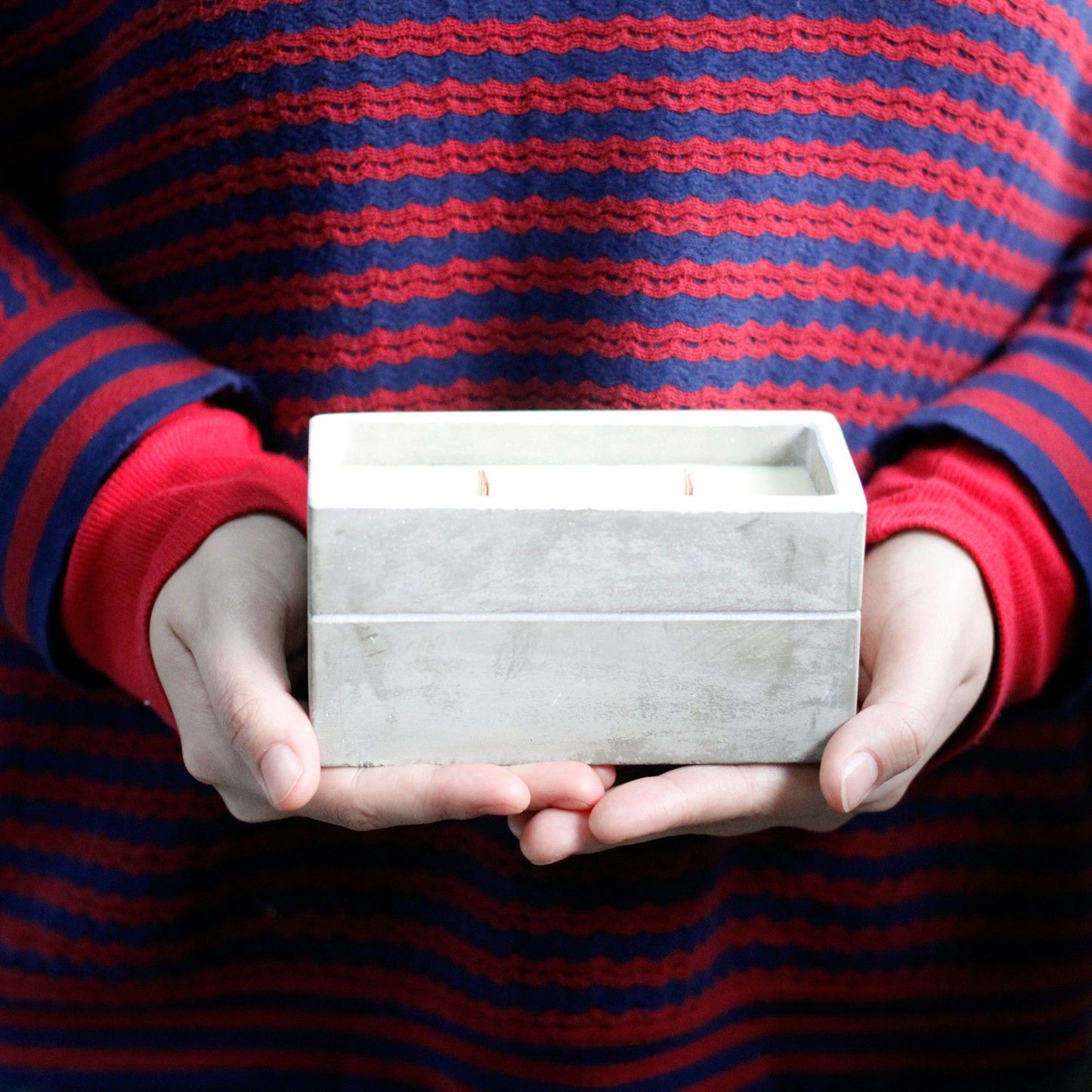 Manos sosteniendo una vela rectangular de hormigón gris con mecha de madera, estilo rústico y minimalista.