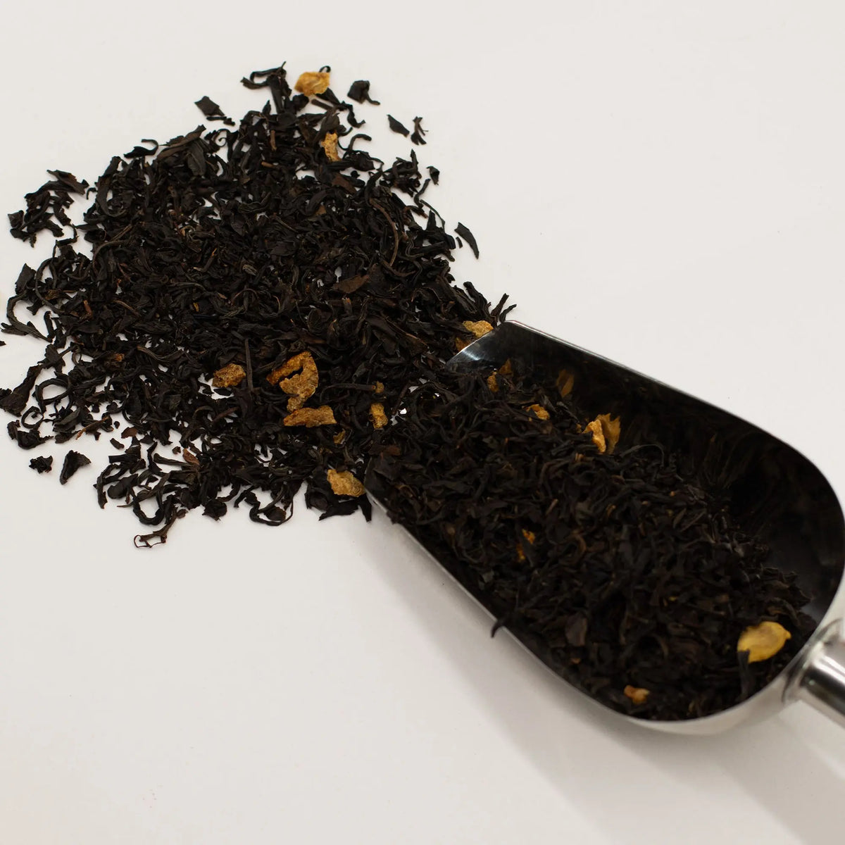 Montón de té negro en hebras con trozos de naranja deshidratada junto a una pala de metal sobre fondo blanco.