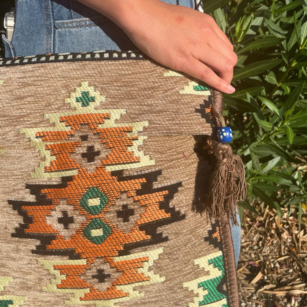 Bolso bandolera de tejido estilo kilim beige con patrones geométricos naranjas, verdes y negros, con borla decorativa.
