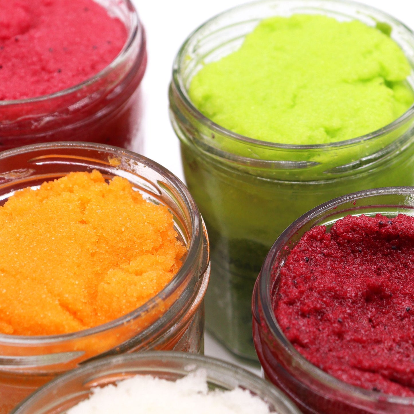 Frascos de vidrio con exfoliantes corporales de azúcar en colores rojo, naranja, verde y blanco sobre fondo blanco.