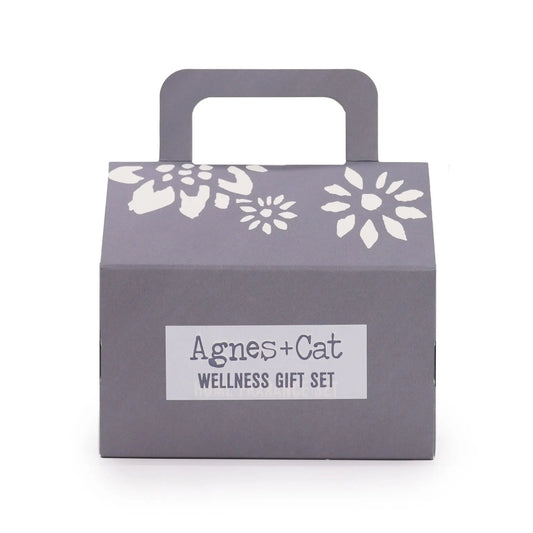 Caja de regalo gris con asa, detalles florales blancos y etiqueta central "Agnes + Cat Wellness Gift Set".