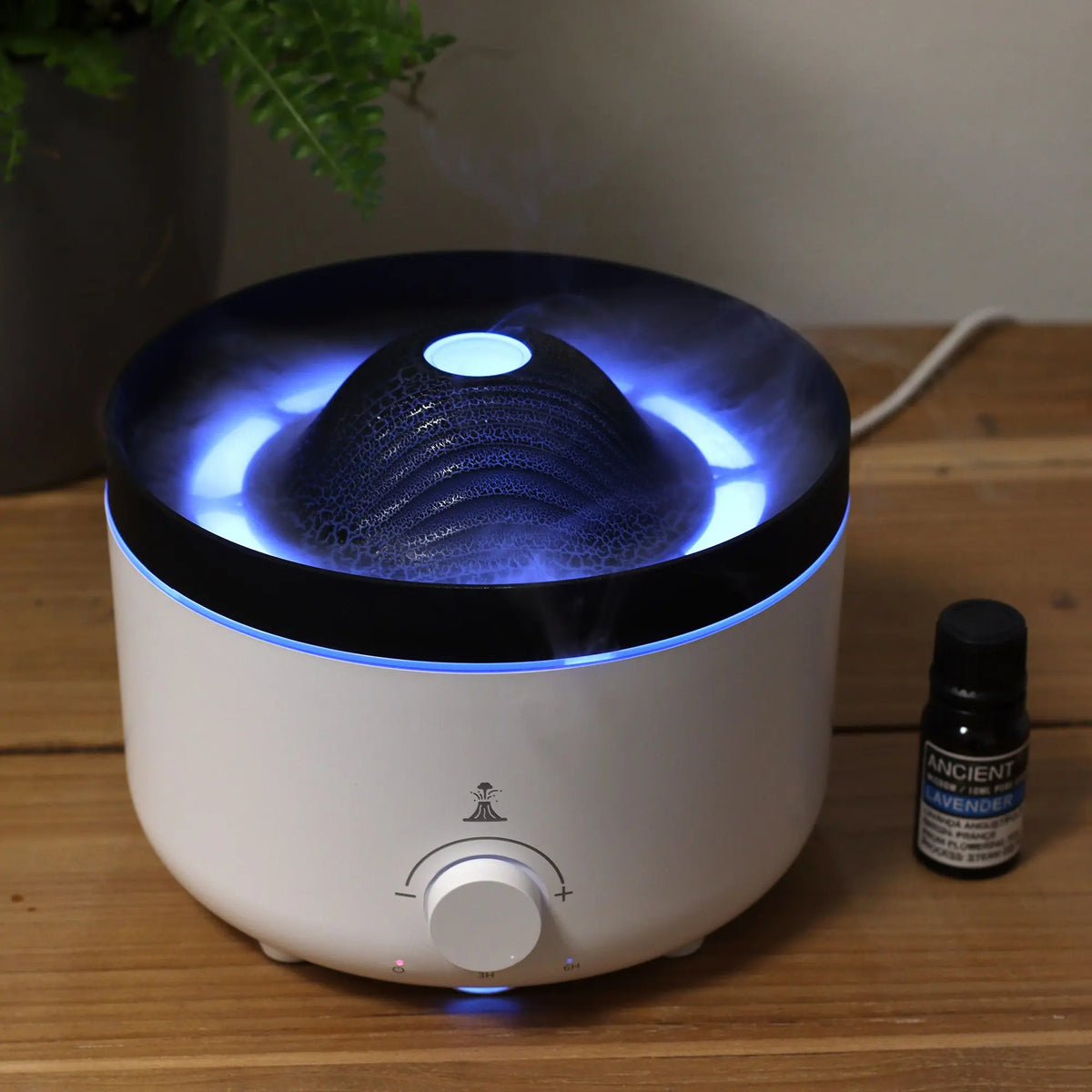 Difusor Ancient Wisdom blanco y negro con efecto volcán, luz LED azul y perilla de control sobre mesa de madera.