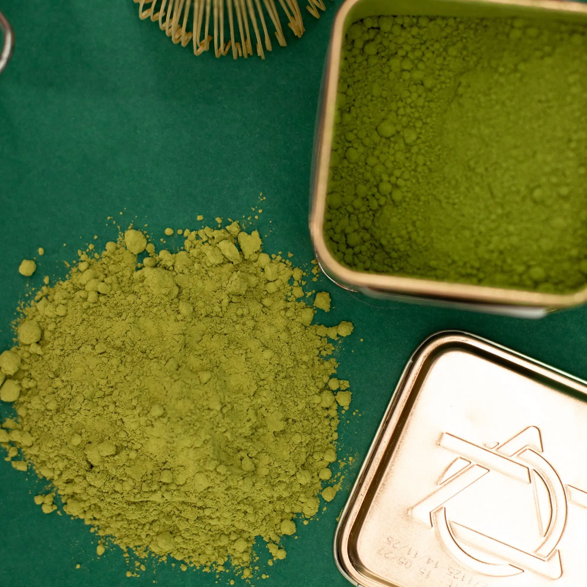 Lata metálica cuadrada abierta con té matcha en polvo, junto a una porción esparcida sobre fondo verde oscuro.