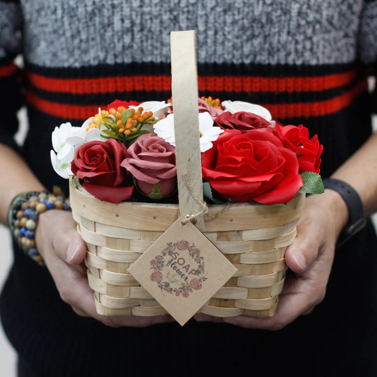 Persona sosteniendo una cesta de mimbre con un ramo de flores de jabón rojas, rosas y blancas.