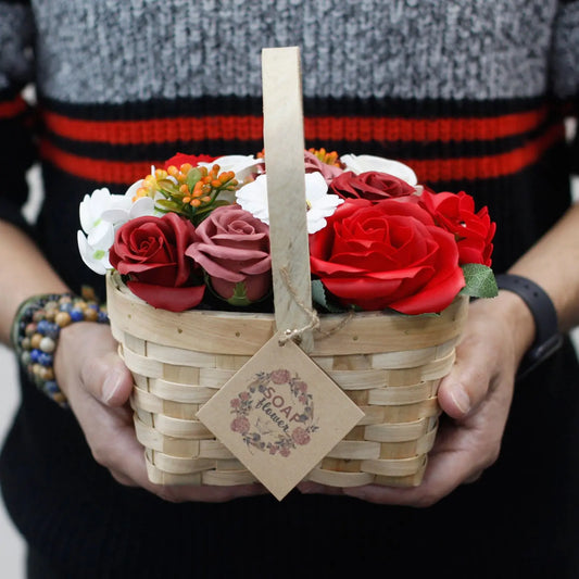 Manos sosteniendo una cesta de mimbre con flores de jabón rojas, rosadas y blancas con una etiqueta de cartón colgada.