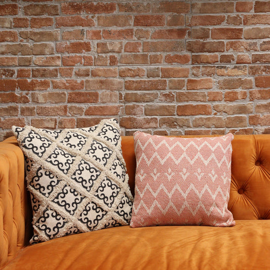 Funda de cojín rosa de 40x40cm con patrón geométrico estilo indio sobre sofá naranja frente a pared de ladrillo.