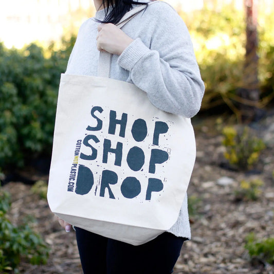 Bolsa de tela de algodón color crudo con el texto "SHOP SHOP DROP" en letras negras grandes, colgada al hombro.