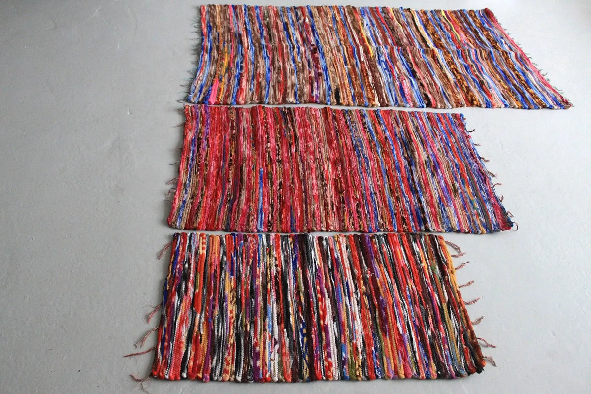 Tres alfombras rectangulares de trapo con franjas verticales multicolor, estilo artesanal indio sobre fondo gris.