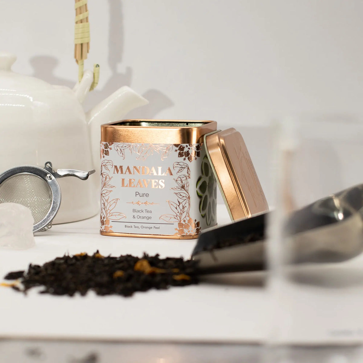 Lata cuadrada de té color blanco y cobre junto a hojas de té negro, un infusor metálico y una tetera sobre fondo claro