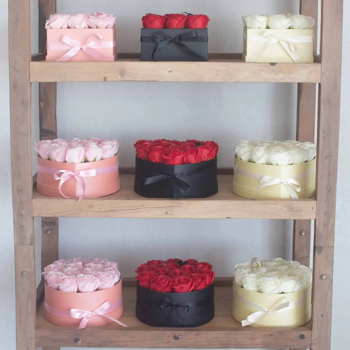Cajas de regalo con rosas de jabón en tonos rosa, rojo y blanco, decoradas con lazos, exhibidas en estantes de madera.