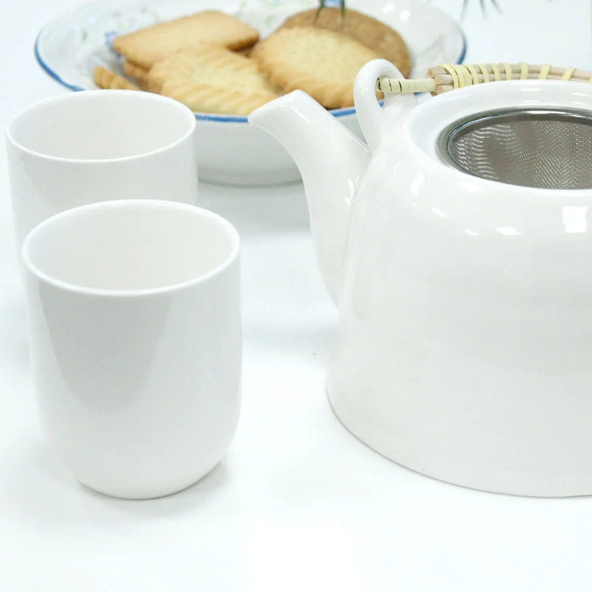 Set de tetera Ancient Wisdom: cerámica blanca, asa de mimbre, filtro metálico y dos tazas a juego con galletas.