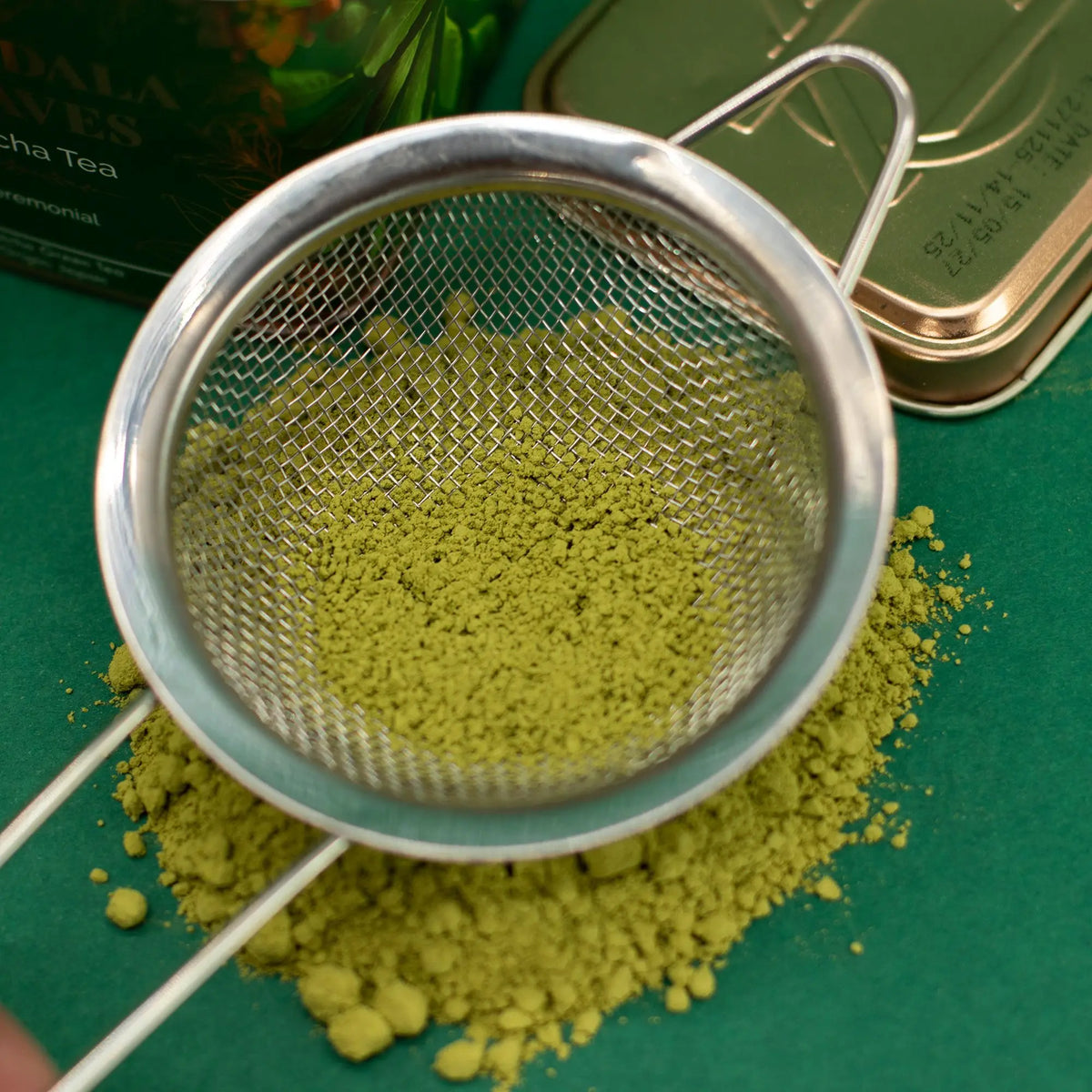 Colador metálico con polvo de té matcha verde vibrante sobre una superficie verde junto a una lata de té dorada