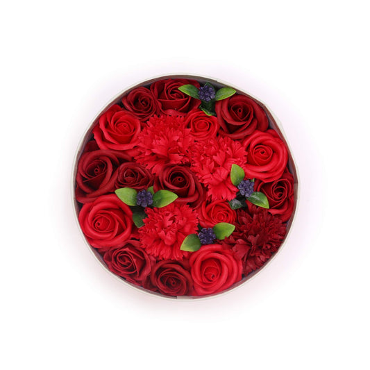 Caja redonda con rosas rojas y claveles de jabón, decorada con hojas verdes y pequeños detalles florales violetas