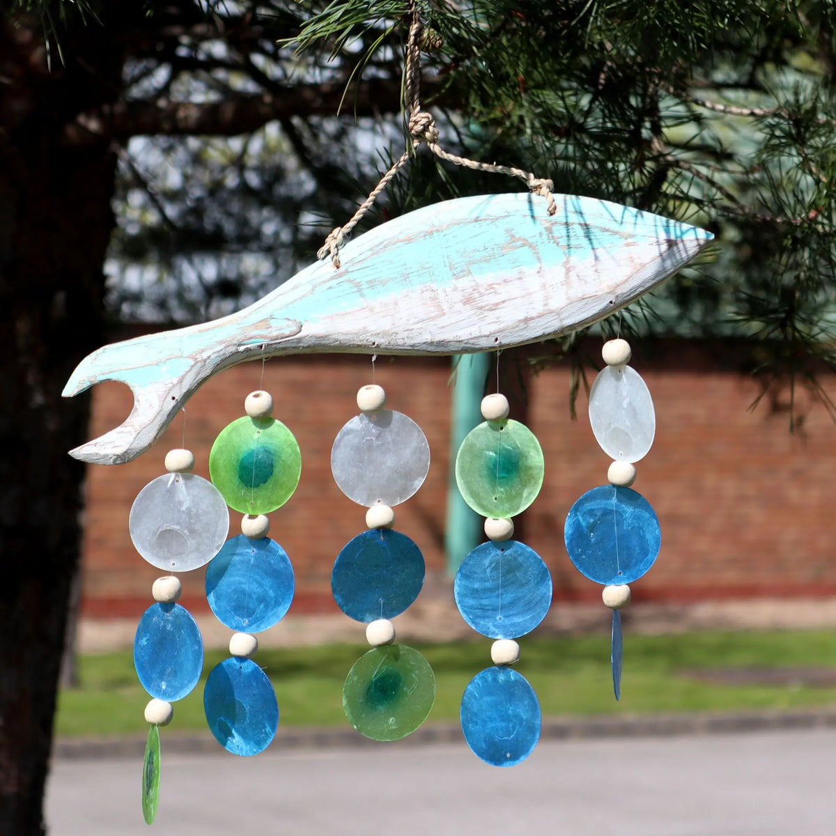 Carillón de madera con forma de pez y círculos colgantes de colores azul y verde tipo gota, colgado en un árbol.