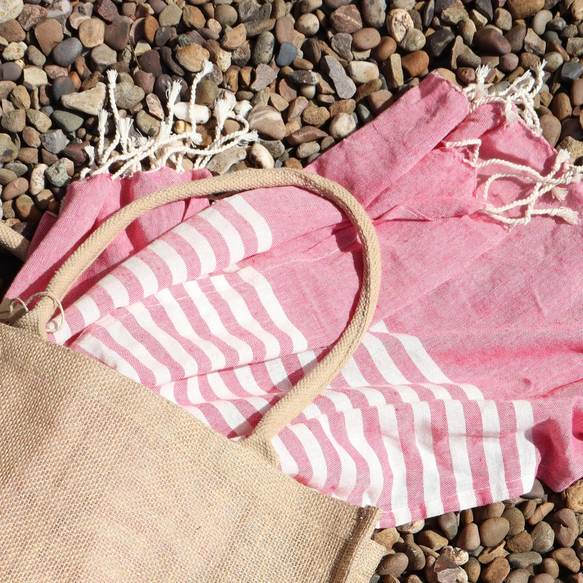 Pareo de algodón rosa con rayas blancas y flecos, junto a un bolso de yute sobre un suelo de piedras.