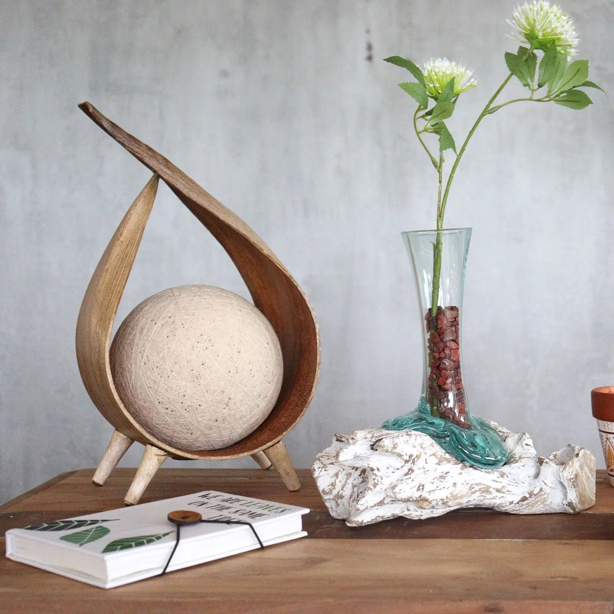 Florero de vidrio fundido sobre base de madera blanqueada, con flor blanca, junto a decoración de madera y libro.