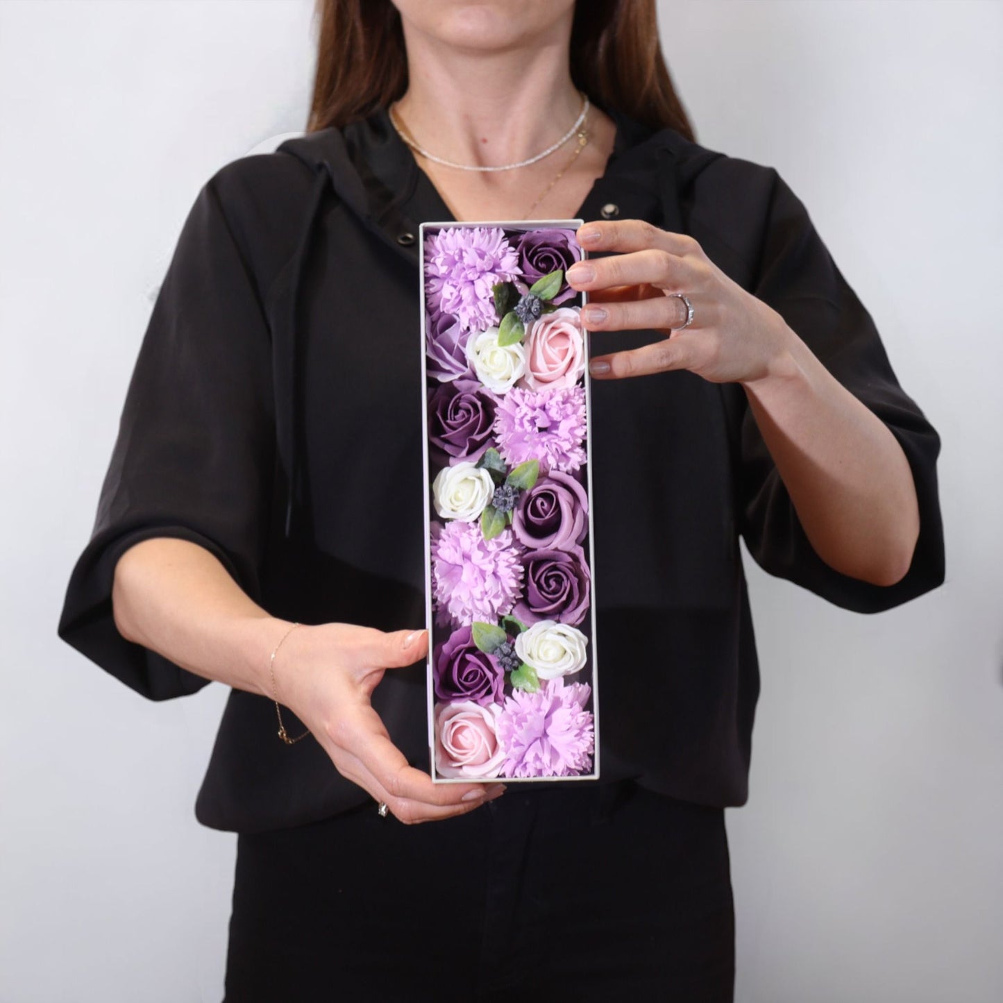Persona sosteniendo una caja larga con rosas y claveles de jabón en tonos lavanda, morado y blanco.