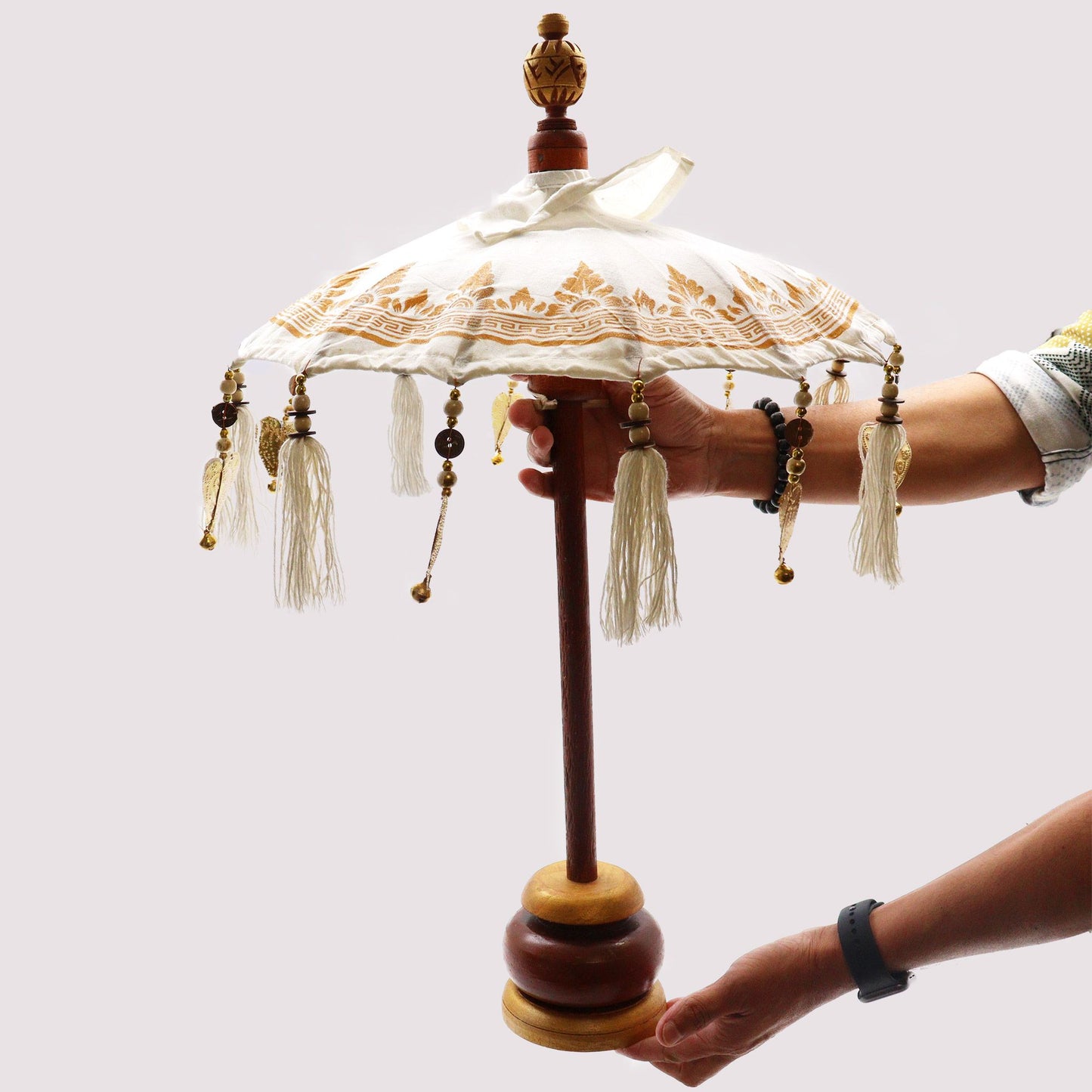 Sombrilla decorativa de lona blanca con detalles dorados, flecos, cuentas y base de madera sostenida por unas manos.