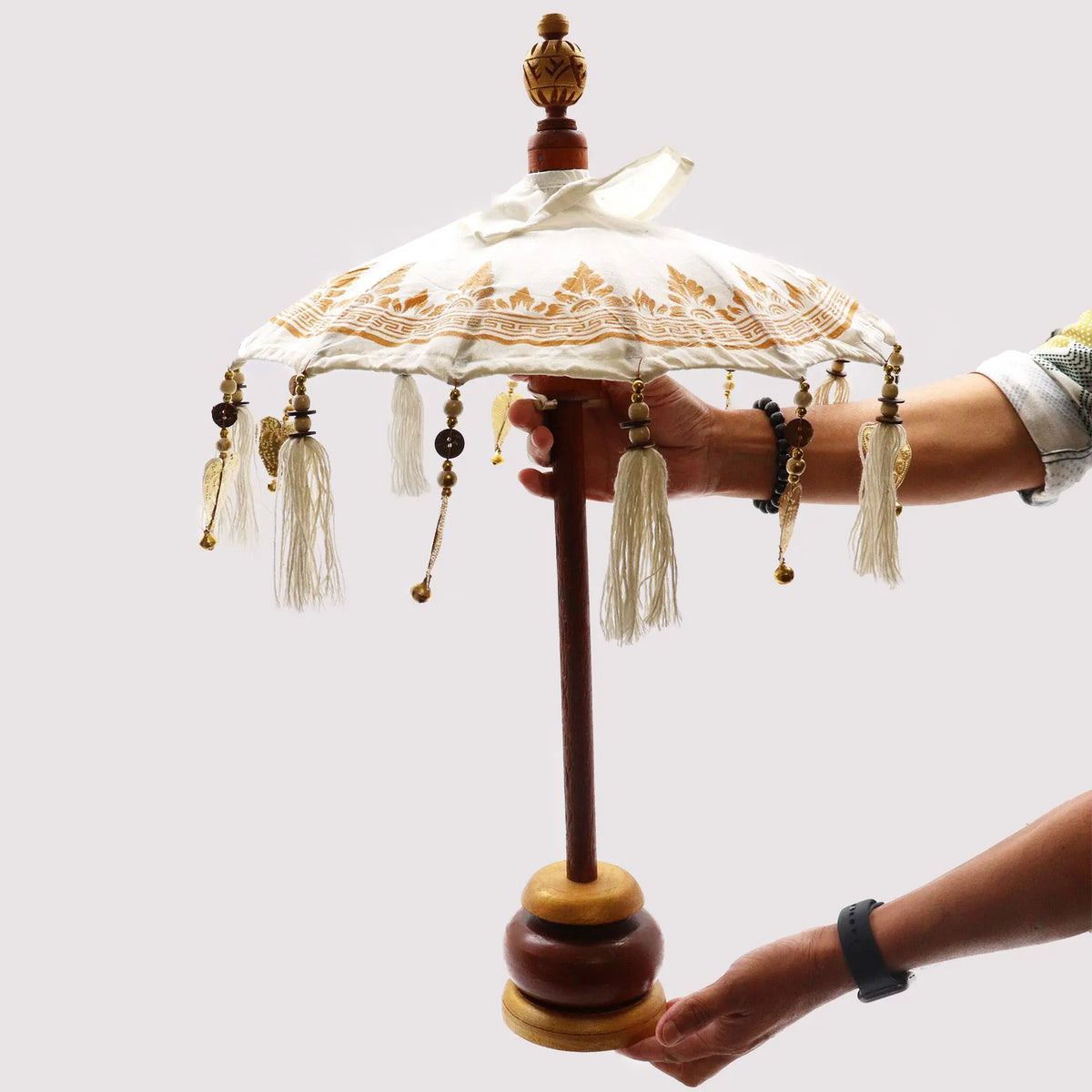 Sombrilla decorativa balinesa de tela blanca con detalles dorados, borlas, cuentas y base de madera sobre fondo blanco.