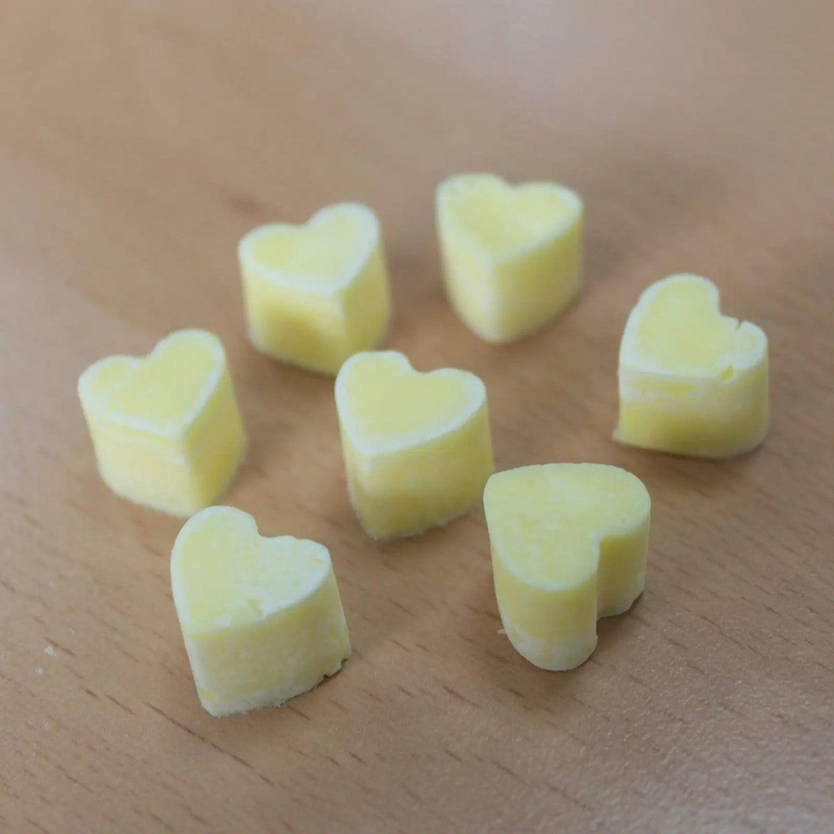 Siete pequeñas piezas de cera aromática en forma de corazón, de color amarillo claro, sobre una superficie de madera.