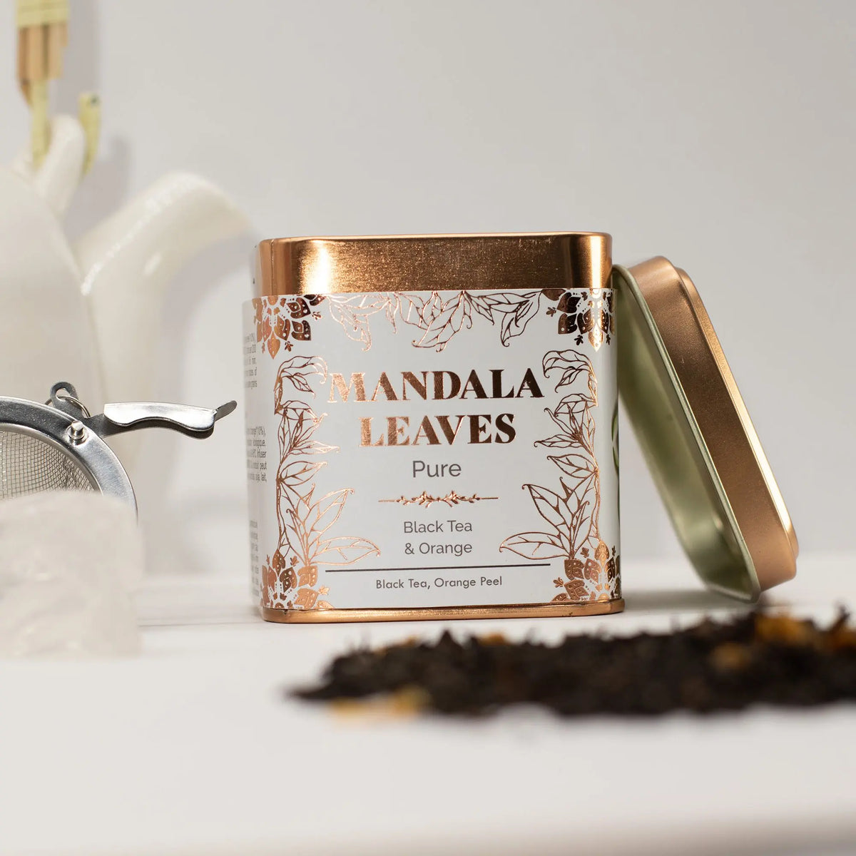 Lata metálica cuadrada de té negro y naranja, con diseño blanco y dorado, junto a hojas de té sobre fondo blanco.