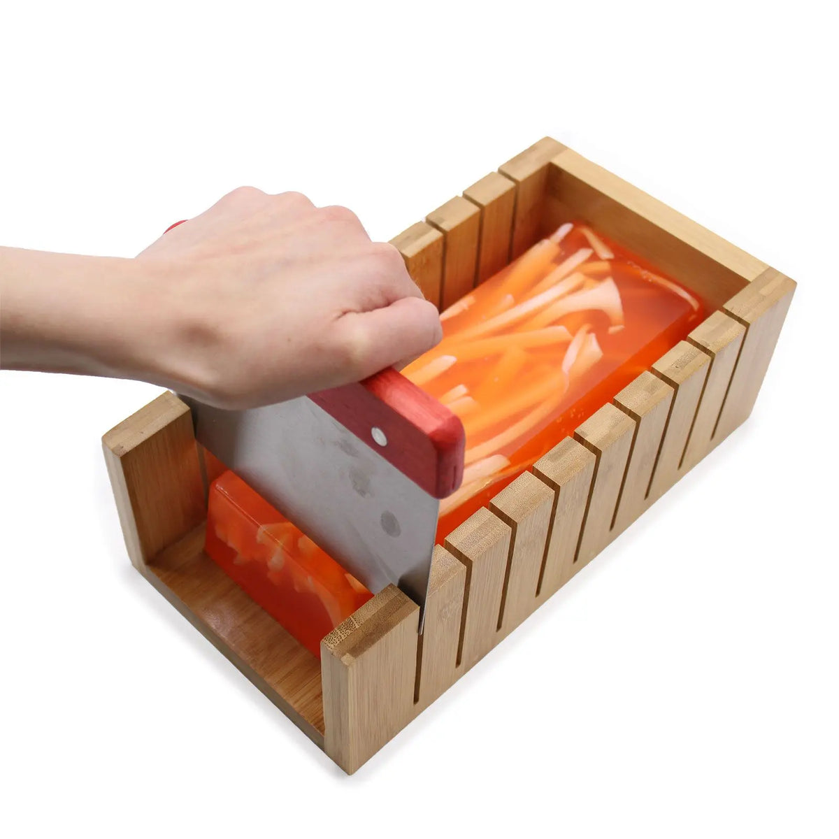 Cortador de madera para jabón con ranuras guía y cuchilla metálica cortando un bloque de jabón naranja traslúcido.