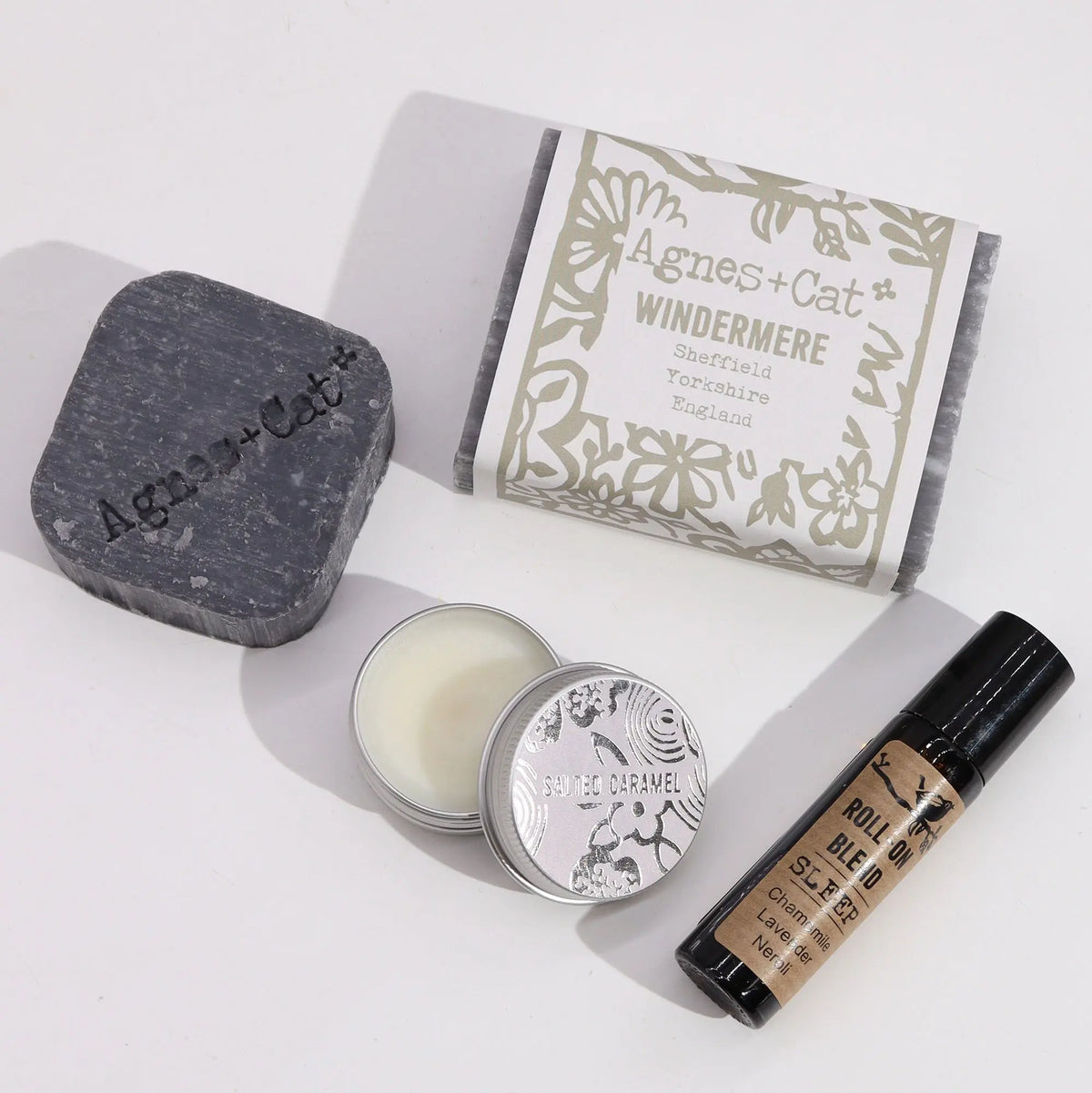Set de regalo: jabón gris, bálsamo labial metálico y roll-on de aromaterapia sobre fondo blanco. Estilo artesanal.