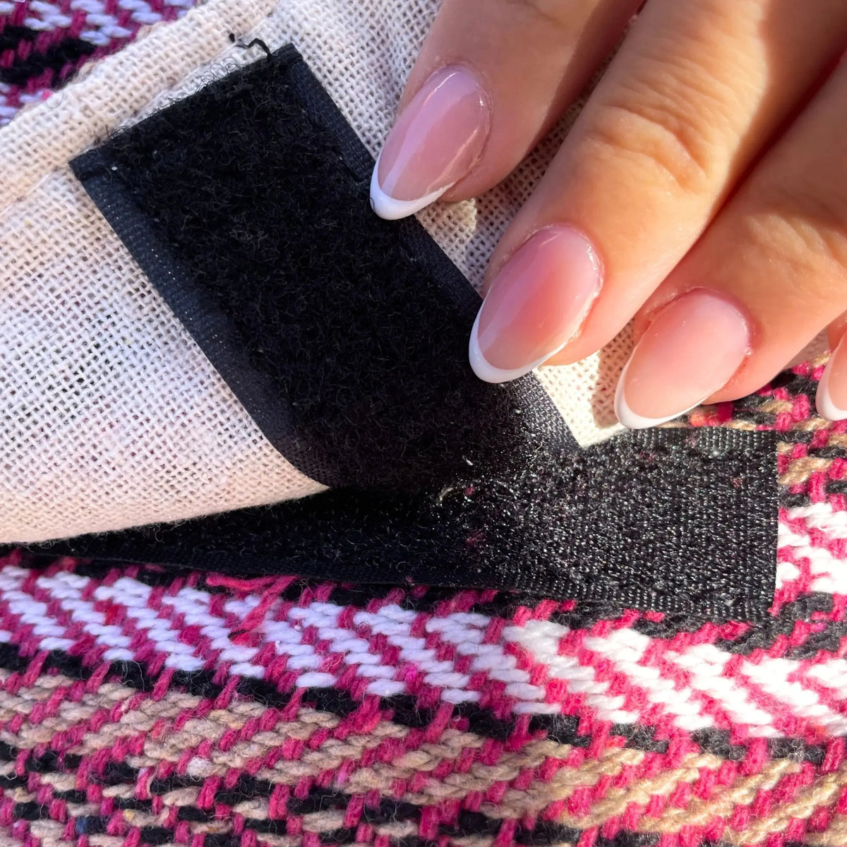 Mano abriendo cierre de velcro negro sobre tela tejida con patrón geométrico en tonos rosa, blanco y beige.