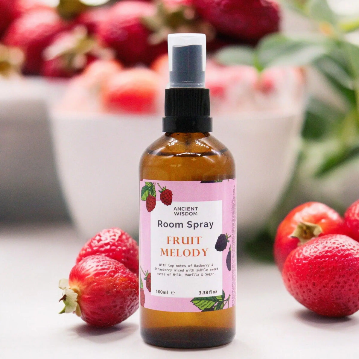 Spray ambientador de 100ml en botella de vidrio ámbar con etiqueta rosa y fresas frescas al fondo sobre mesa blanca.