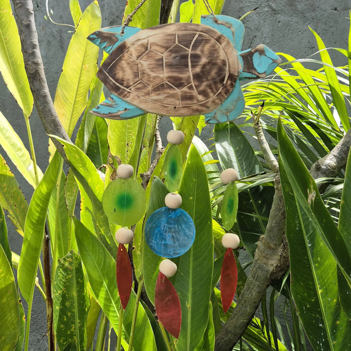 Carillón de madera con forma de tortuga, tonos aqua y adornos colgantes de colores, suspendido entre plantas verdes.