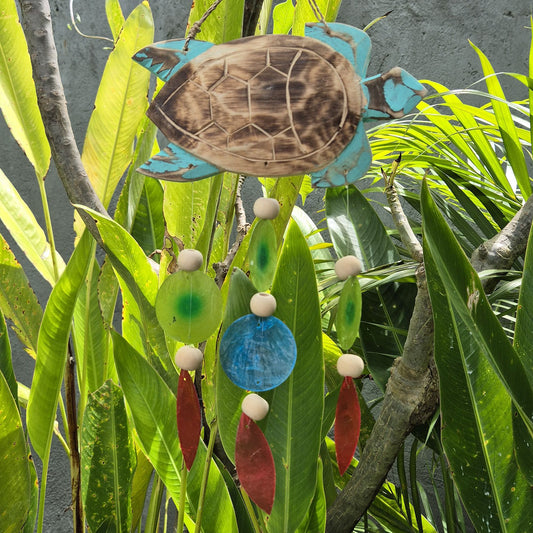 Carillón de madera con forma de tortuga, tonos aqua y adornos colgantes de colores, suspendido entre plantas verdes.