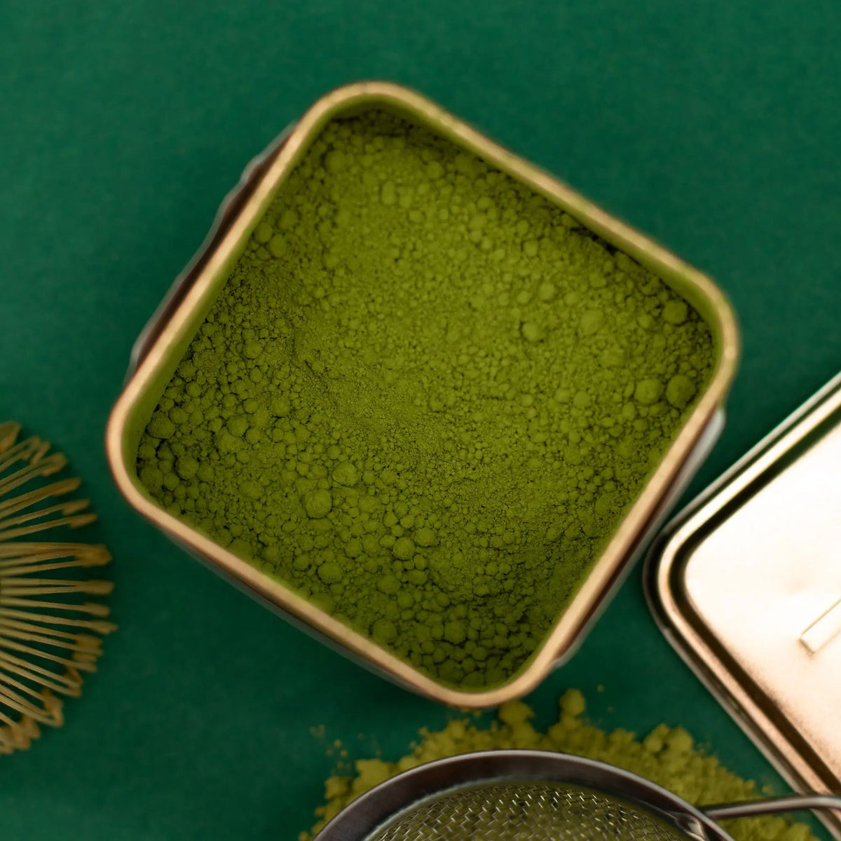 Lata cuadrada abierta con té matcha en polvo, batidor de bambú y colador sobre un fondo verde.