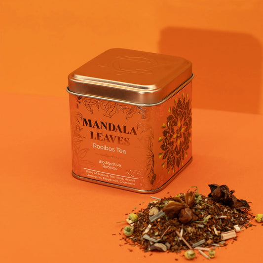Lata cuadrada de color cobre con grabados florales, junto a una mezcla de té rooibos sobre un fondo naranja.