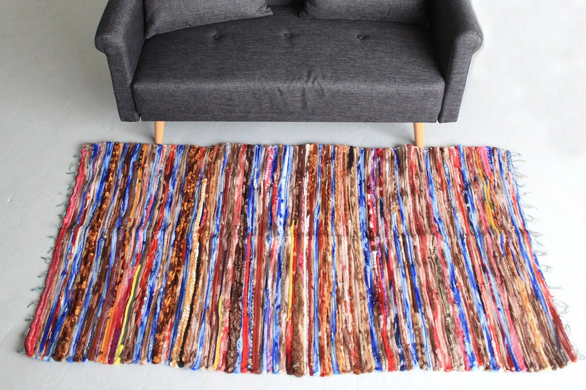 Alfombra de trapo rectangular (105x185cm) con rayas multicolores texturizadas frente a un sofá gris. Estilo artesanal.