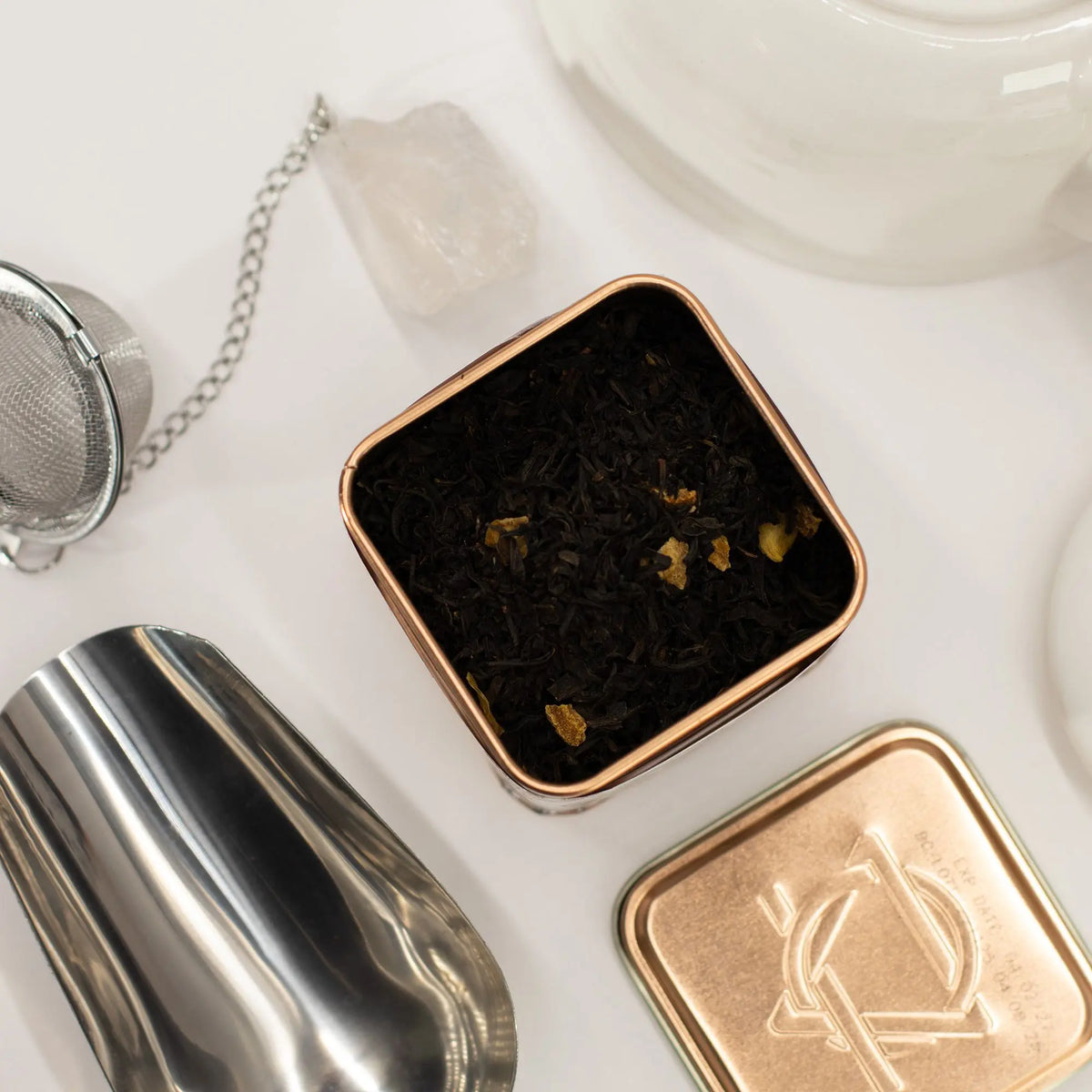 Lata cuadrada con té negro y trozos de naranja junto a cuchara de metal y filtro sobre fondo blanco minimalista