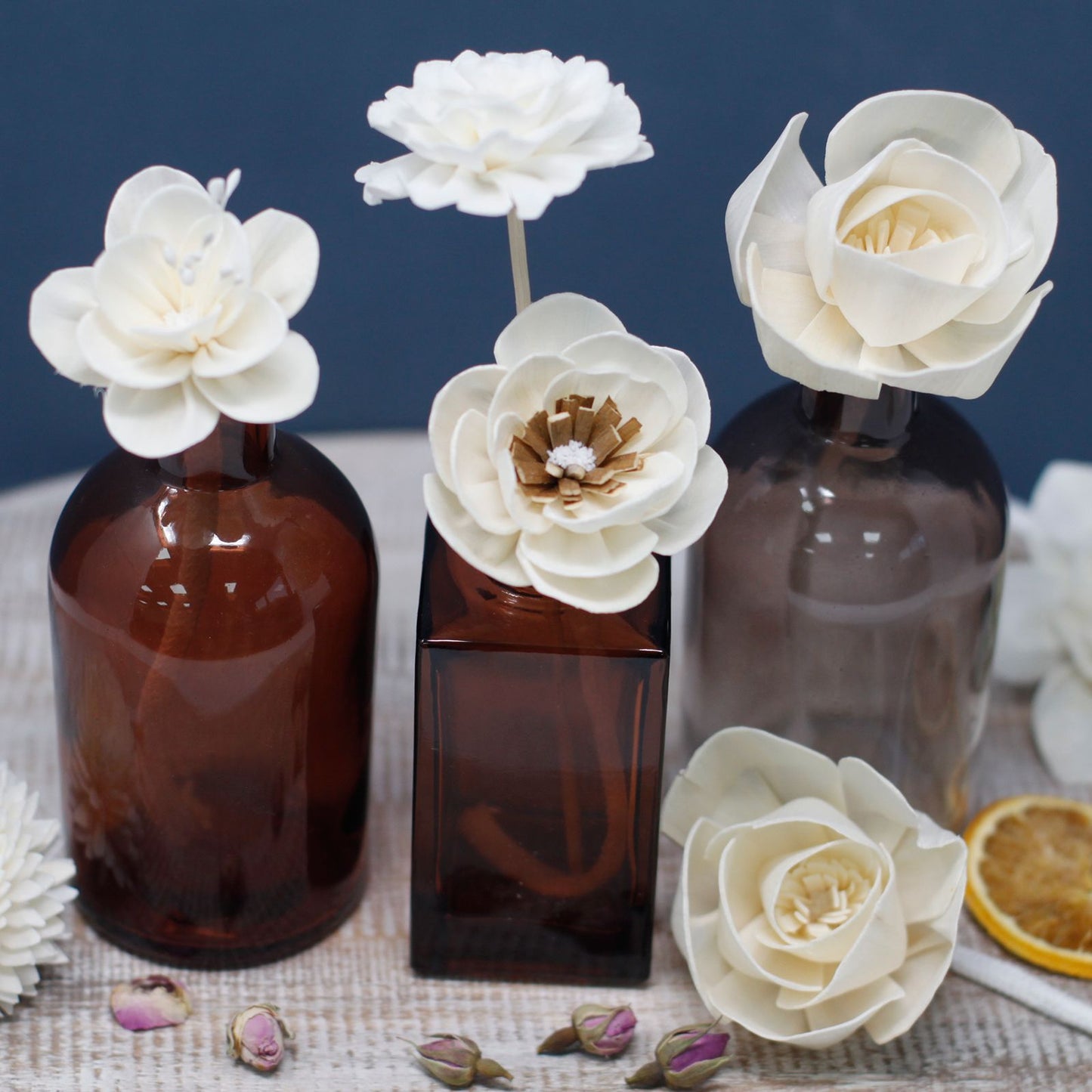 Flores difusoras blancas de madera sobre frascos de vidrio ámbar, decoradas con capullos y rodajas de fruta seca.