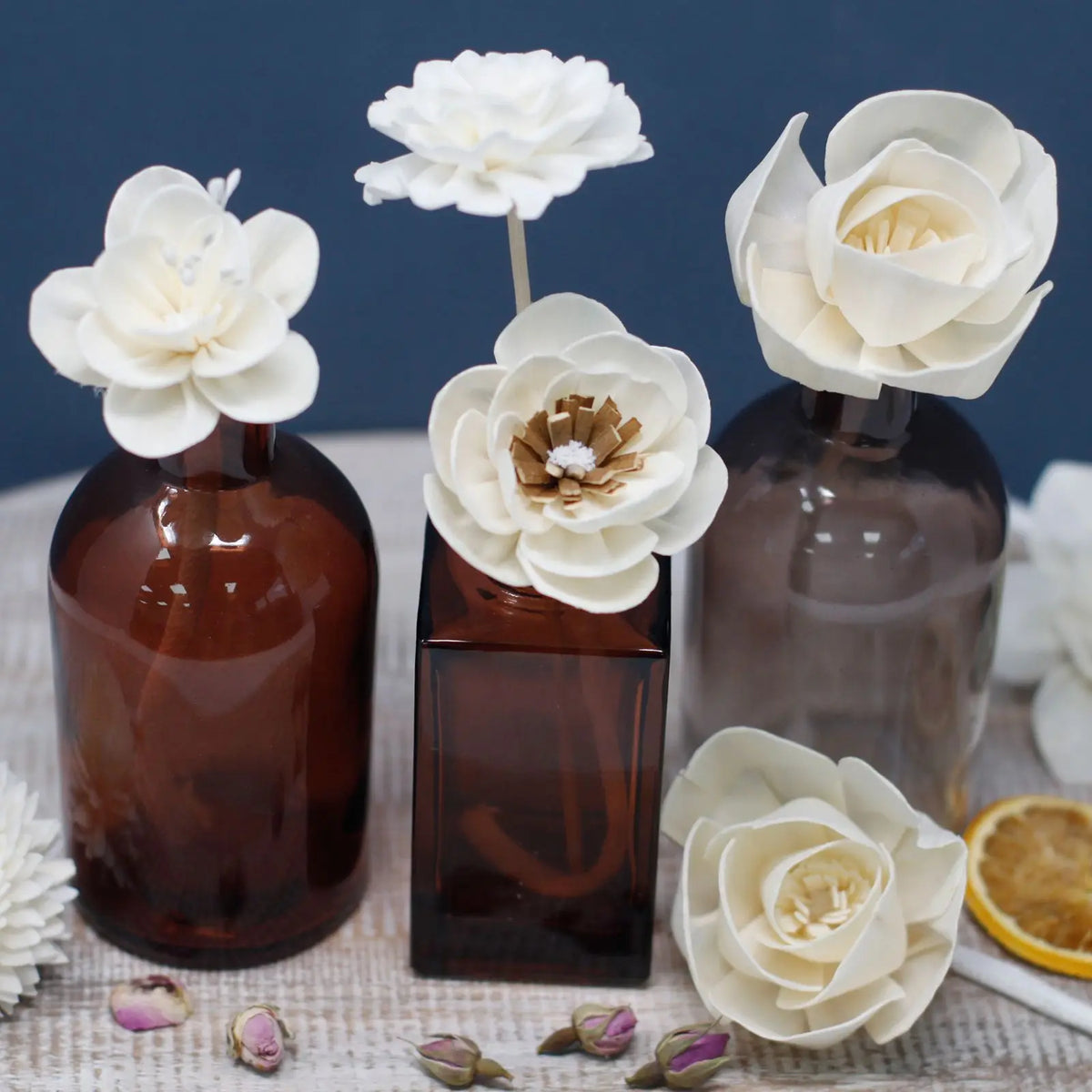 Tres flores de madera blanca sobre botellas de vidrio ámbar, decoradas con capullos de rosa y una rodaja de limón.